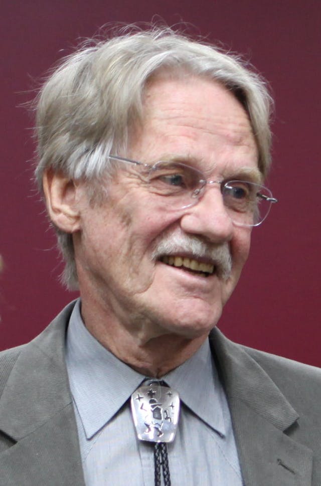 Man with gray hair smiling and wearing glasses. He is dressed in a gray suit and tie with a unique, silver ornament, against a plain burgundy background.