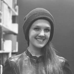 Smiling person wearing a beanie and a leather jacket, standing in front of shelves filled with books in a dimly lit room.