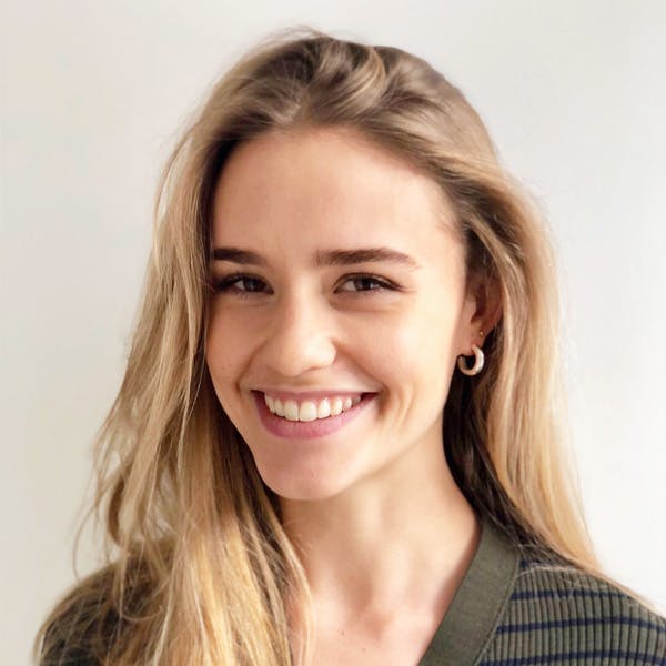 A woman with long blonde hair smiles warmly, wearing a green striped shirt and hoop earrings. She is positioned against a plain white background.