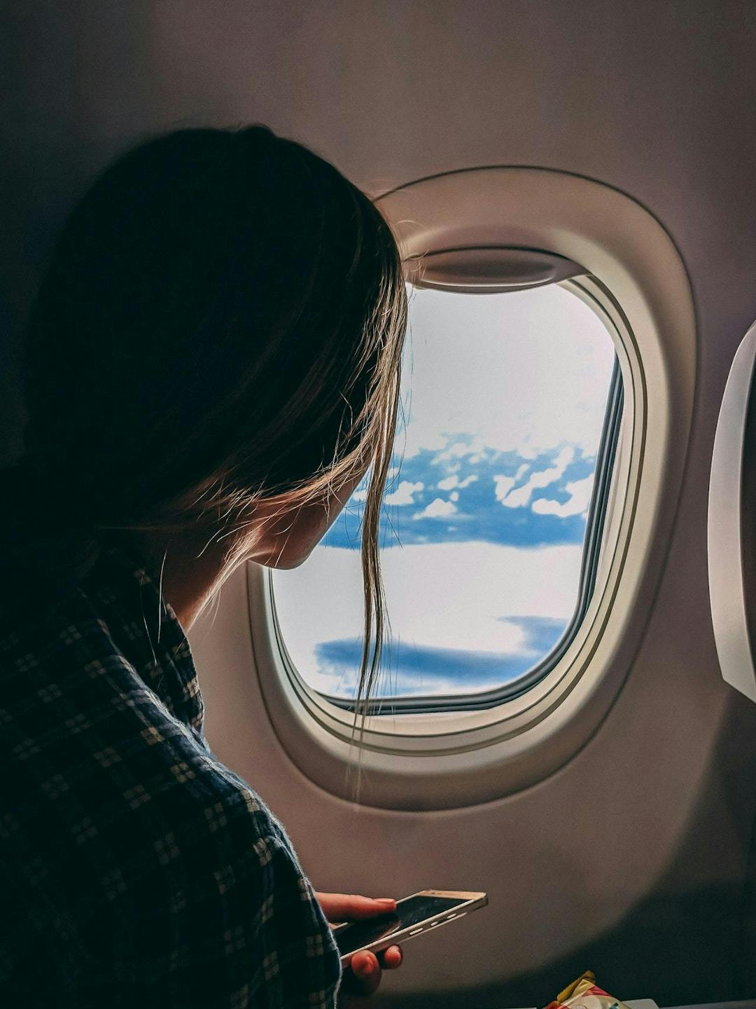 A person holding a phone gazes through an airplane window at clouds and blue sky, creating a serene and contemplative atmosphere.