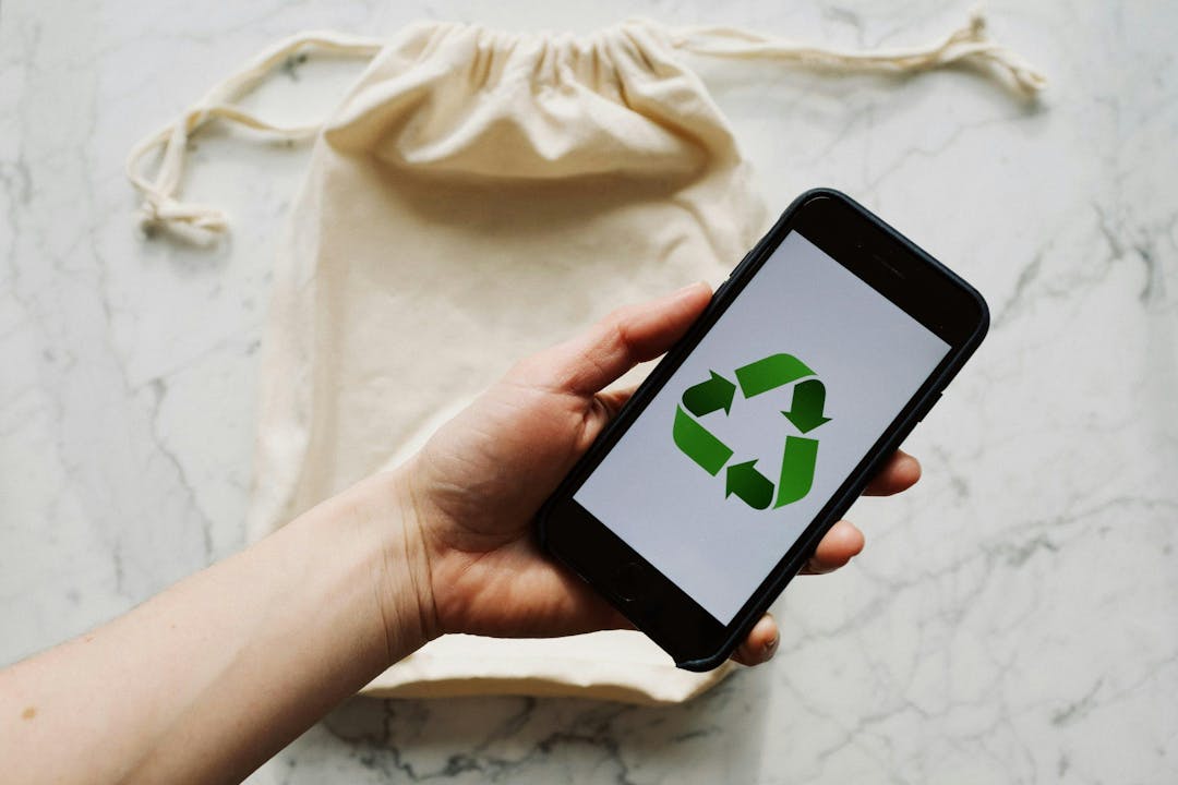 A hand holds a smartphone displaying a green recycling symbol above a beige drawstring bag on a marble surface.