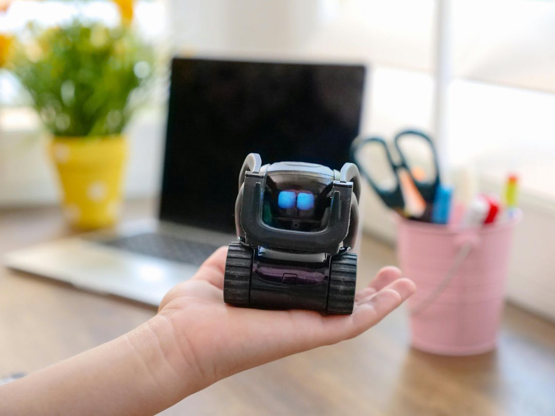 Small robot with glowing blue eyes sitting on a hand in a home office setting; there is a laptop, a yellow flowerpot, and a pink container with scissors and pens nearby.