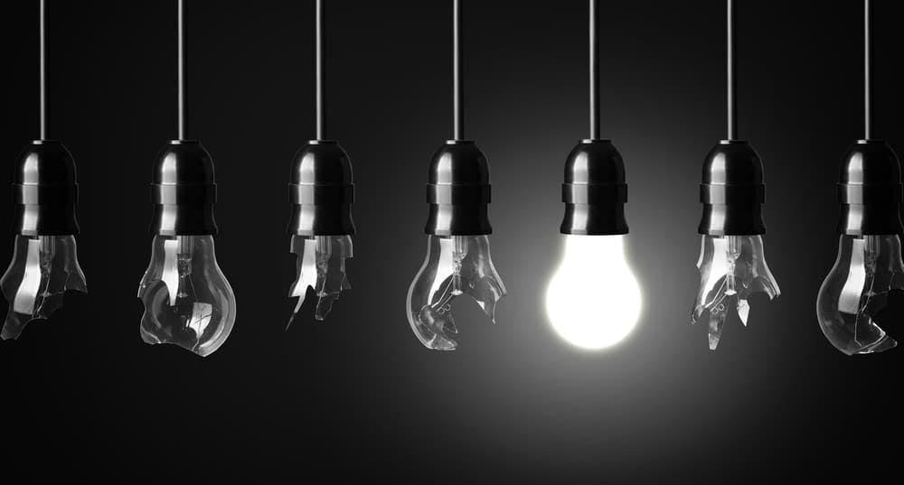 A row of seven hanging light bulbs, six shattered and one intact and glowing, contrasting against a dark background.