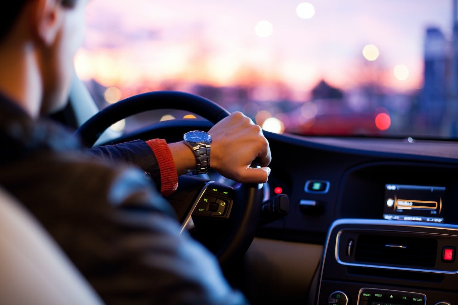 A person is driving a car at dusk, with one hand on the steering wheel, wearing a wristwatch, illuminated dashboard, and blurred city lights visible outside the windshield.