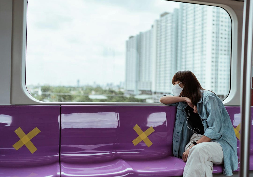 A person wearing a mask and blue jacket is leaning against a window, sitting on a purple train seat with yellow X markers. Outside, high-rise buildings are visible.