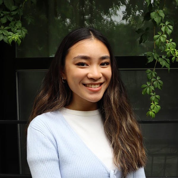 Woman smiles, wearing a light blue sweater over a white shirt. The background shows a dark window surrounded by green leafy plants.