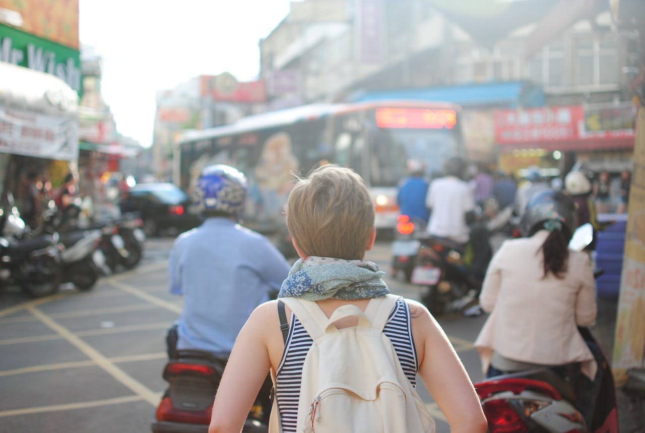 Person with a backpack walking through a busy street, surrounded by motorcycle riders, with a bus and various storefronts visible in the background.