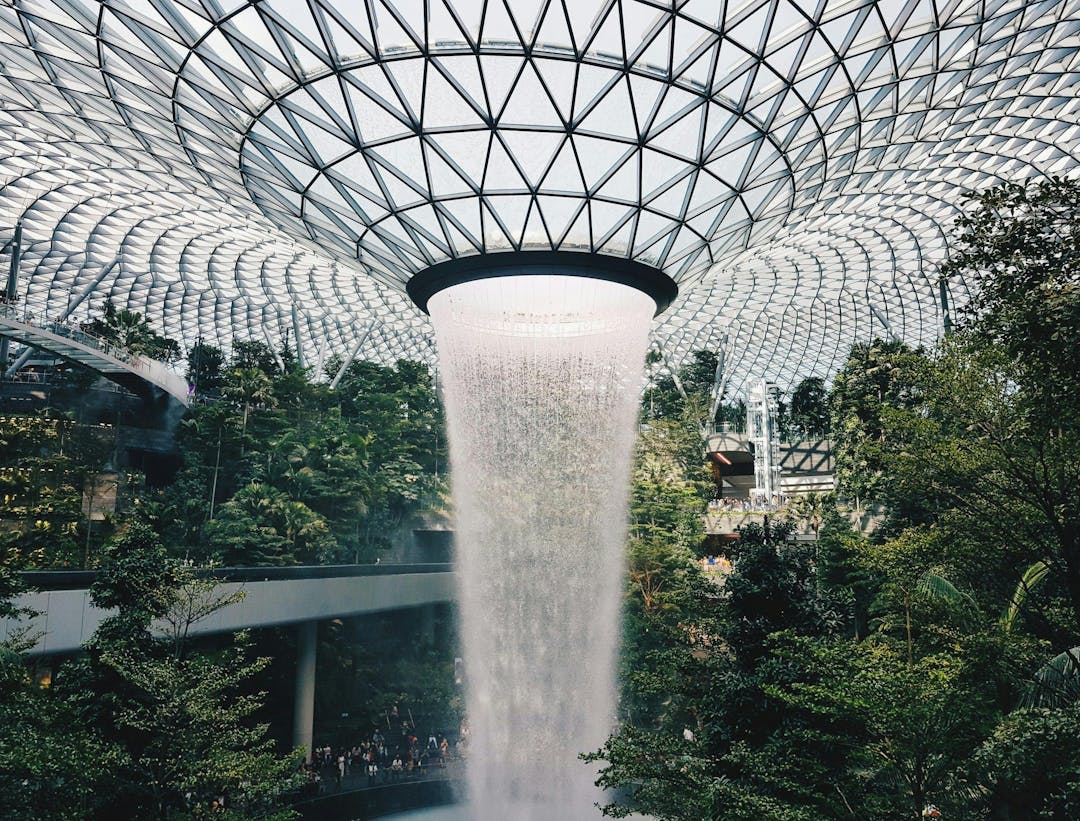 A large, circular indoor waterfall cascades from a glass-domed roof into a lush, verdant garden filled with trees and plants, surrounded by architectural walkways and numerous visitors.