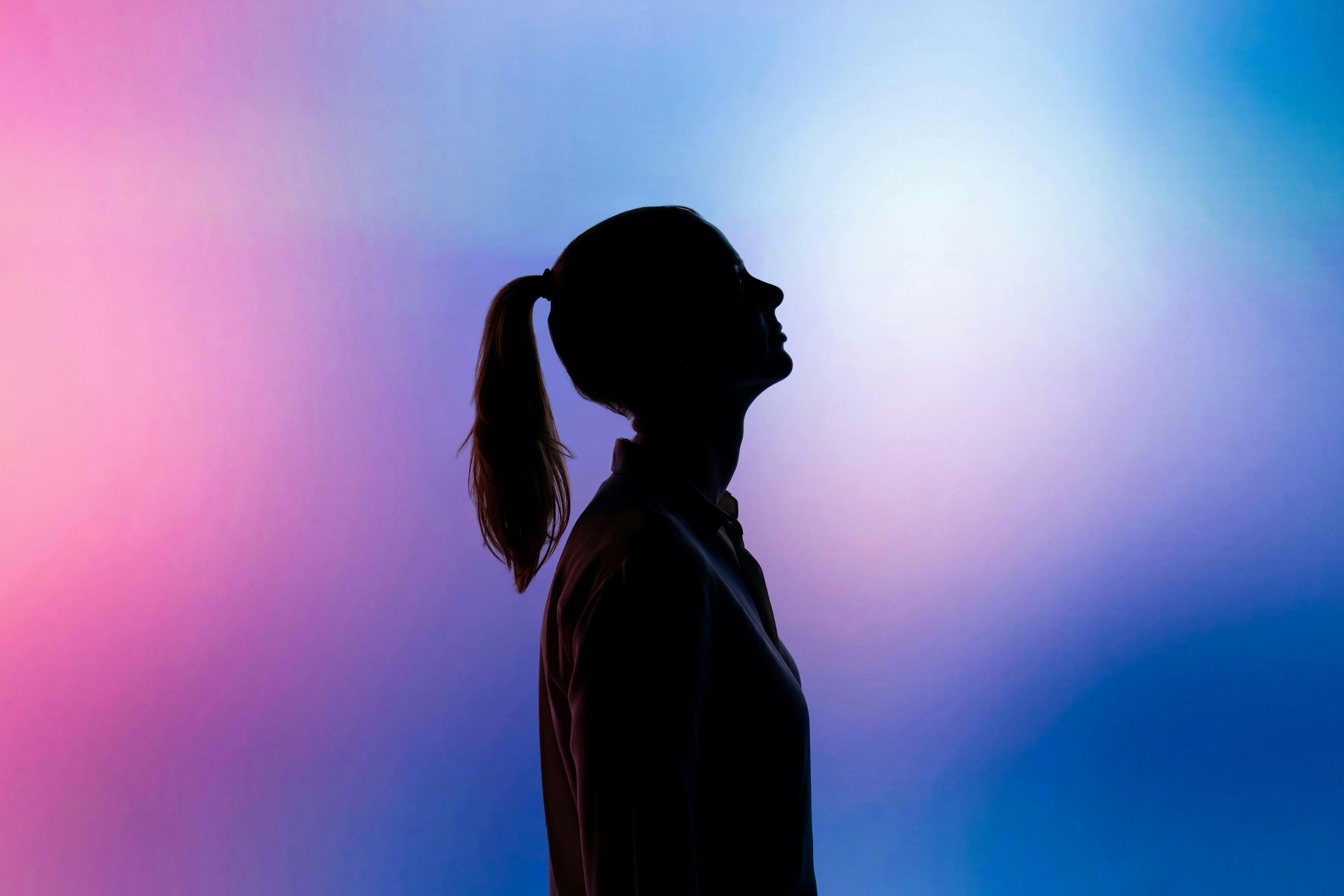 Silhouetted person stands in profile with a ponytail against a vibrant, gradient background of pink, purple, and blue hues.