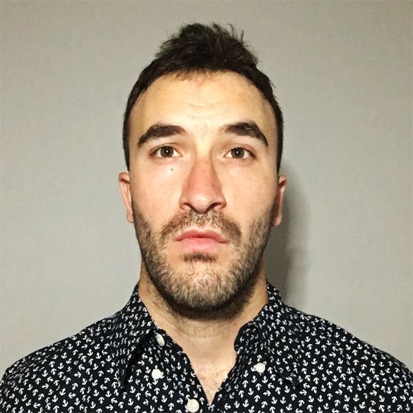 A man with a short beard is staring directly ahead, wearing a dark patterned shirt with white anchors, and standing against a plain gray background.