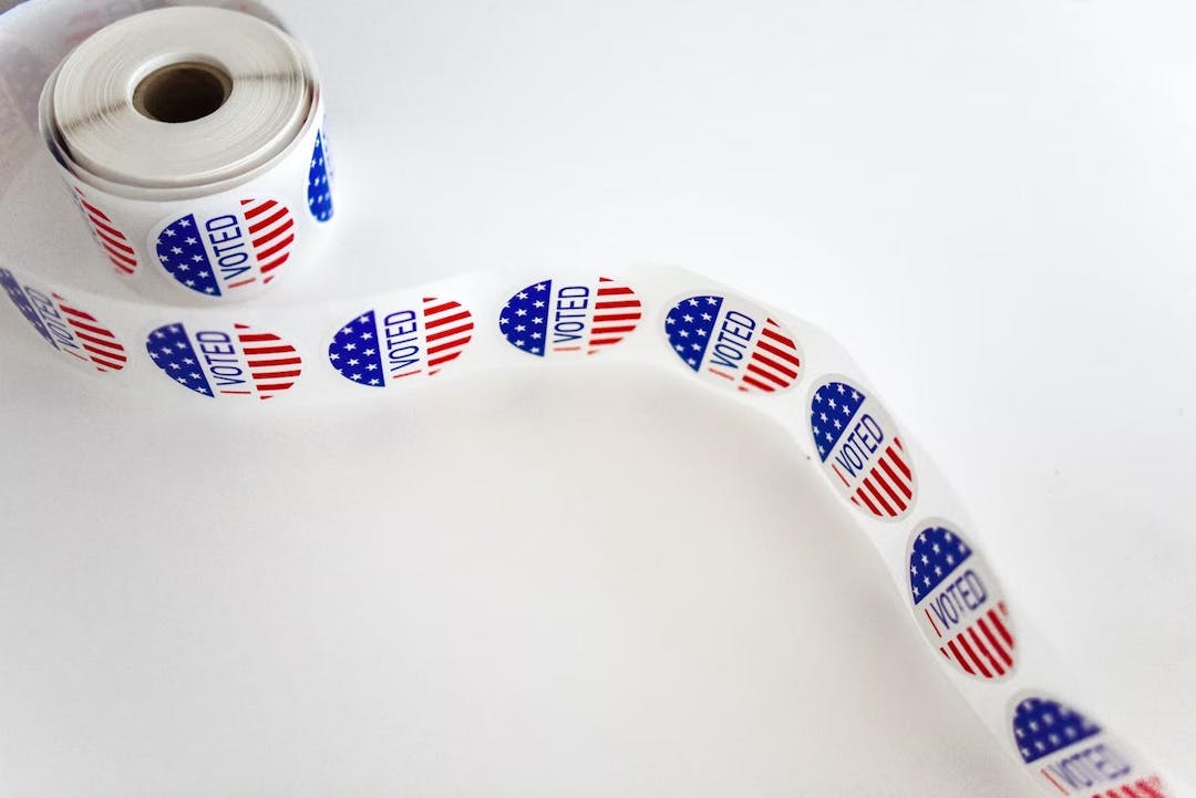 A roll of circular "I Voted" stickers, featuring the American flag's stars and stripes, lies unrolled in a loop on a white surface.