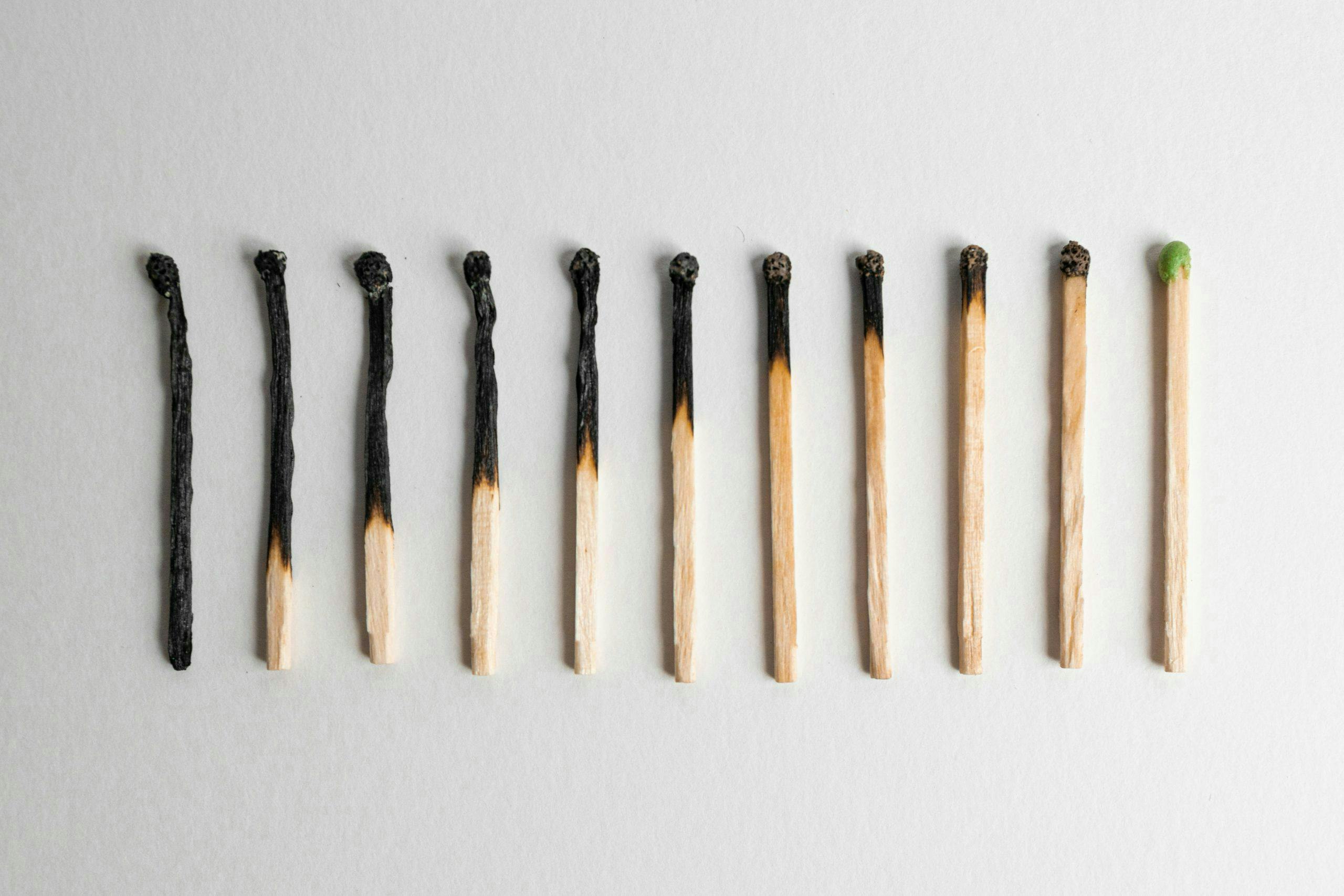 Eleven matches arranged sequentially; ten are burned to varying degrees, and one is unlit with a green tip. They are positioned horizontally on a light grey surface.