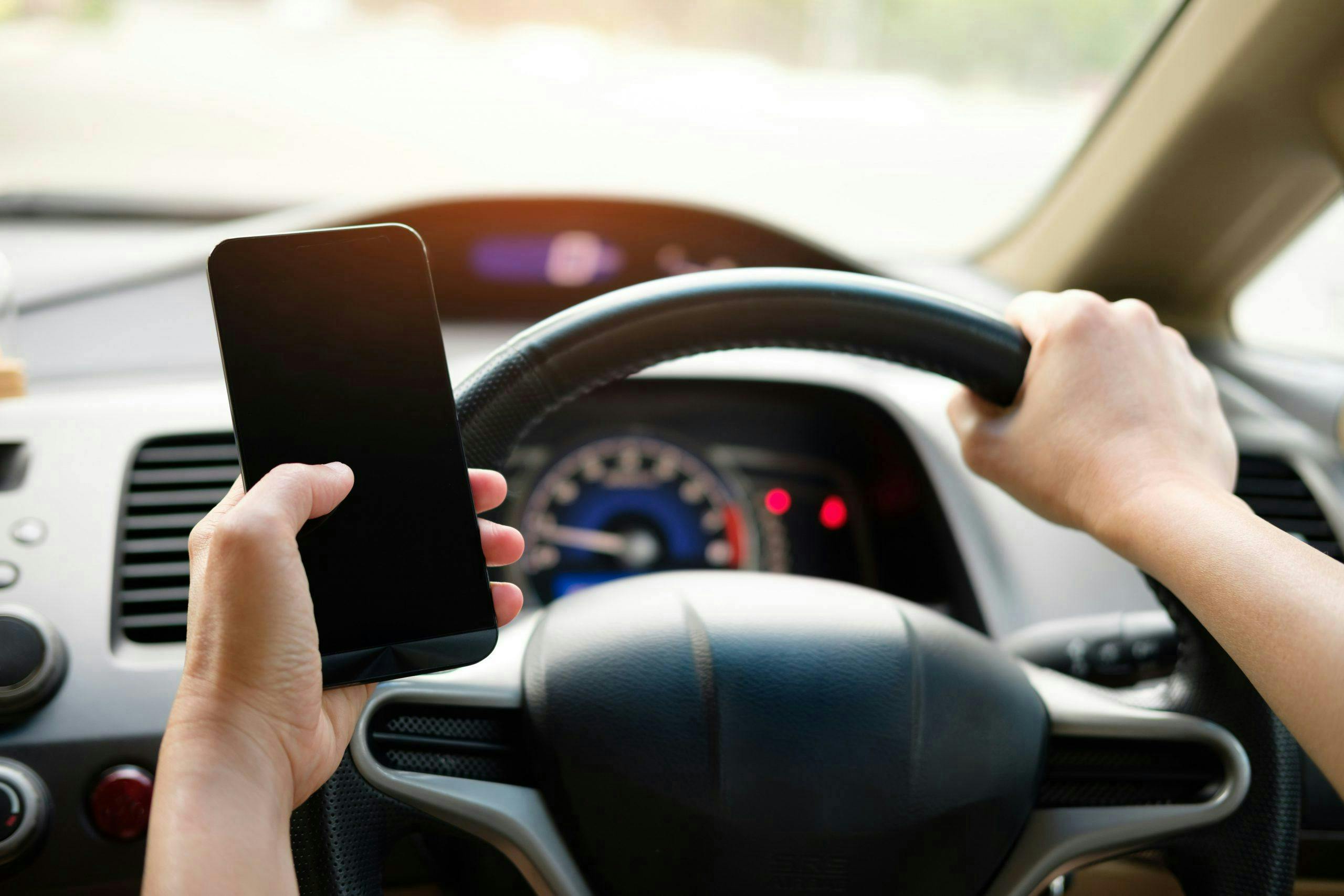 A person holds a smartphone in their left hand and grips a car steering wheel with their right hand, with a dashboard displaying speed and controls in the background.