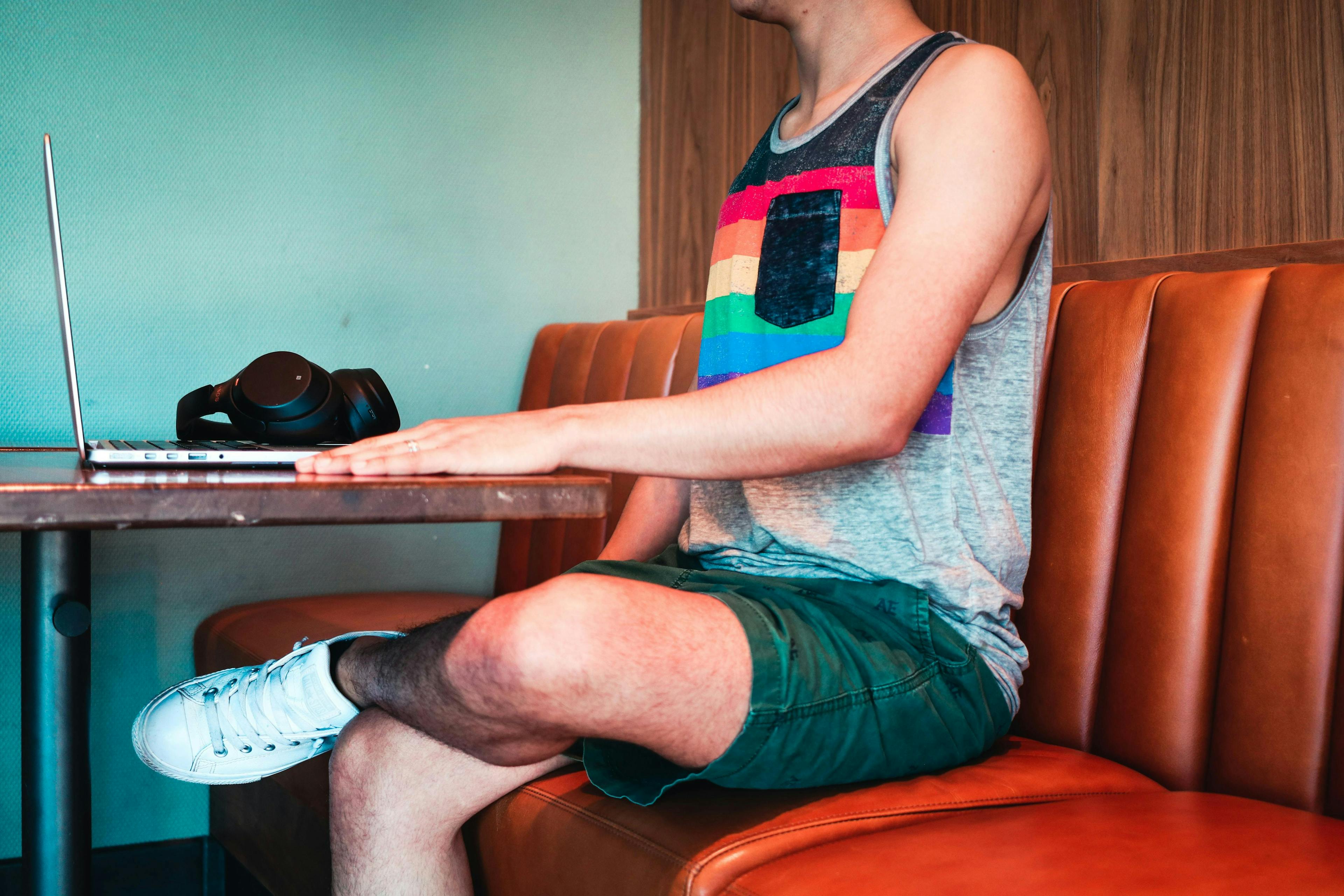 Person wearing a rainbow tank top and green shorts sits at a booth, using a laptop on a wooden table, with black headphones next to the laptop in a cozy indoor setting.