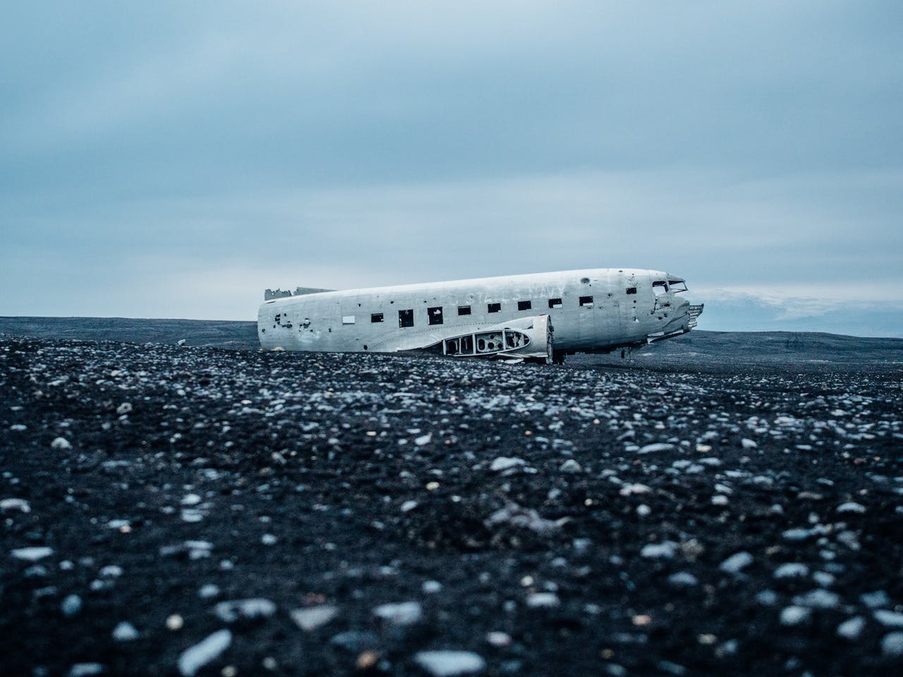 A crashed, weathered airplane lies abandoned on a desolate, rocky beach under an overcast sky.
