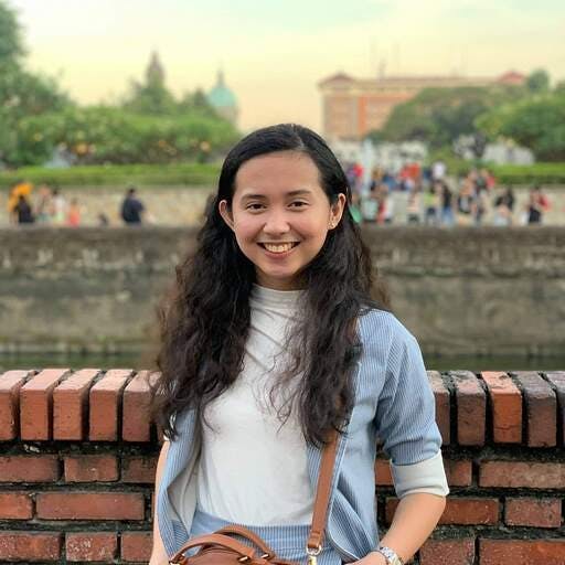 A woman stands smiling, leaning against a brick wall, with lush greenery and a distant historical building in the background. She wears a light blue jacket over a white top.