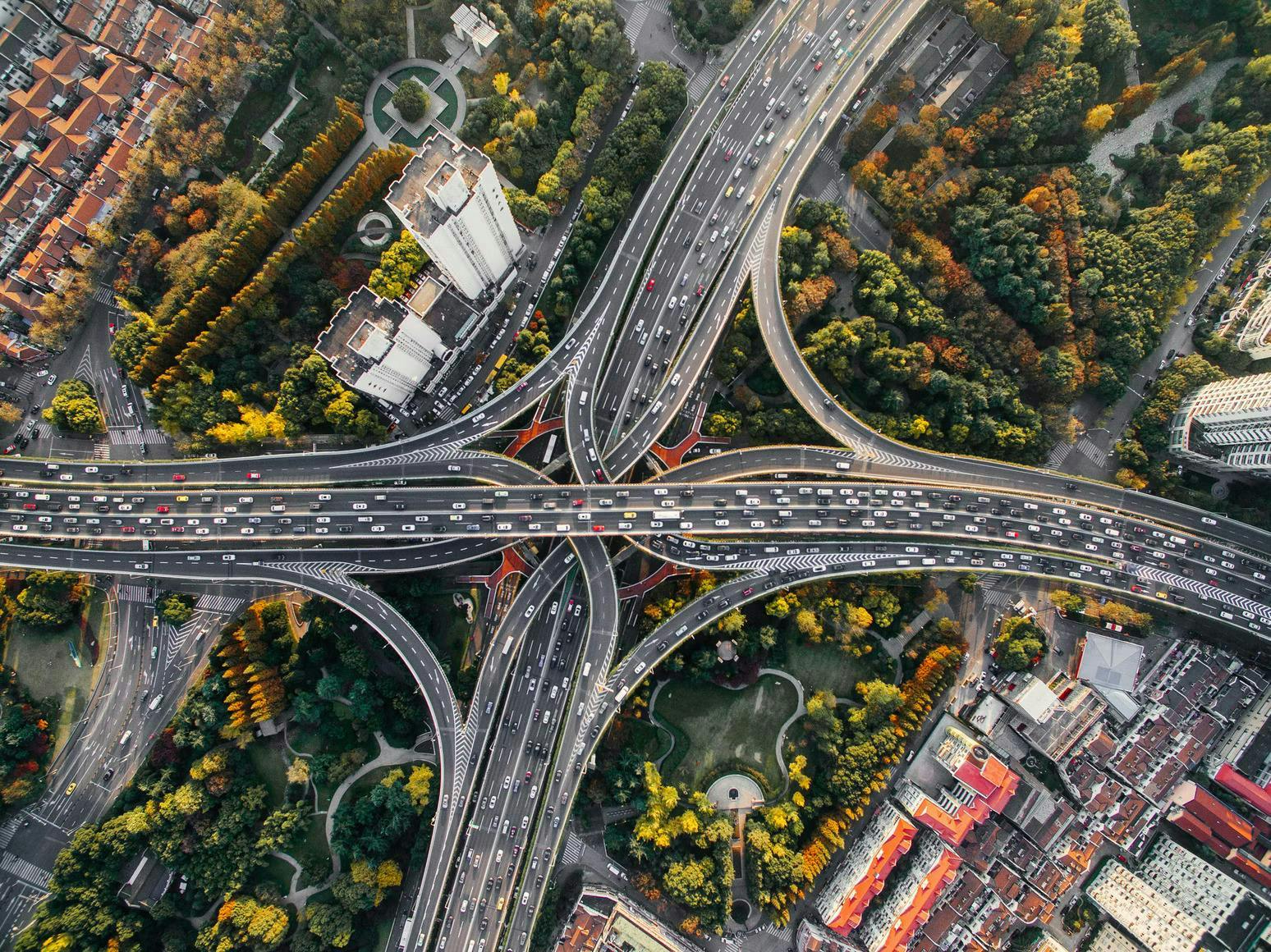 Aerial view of a sprawling, multi-layered highway interchange bustling with dense traffic amidst a mix of urban buildings and lush green spaces adorned with autumn foliage.