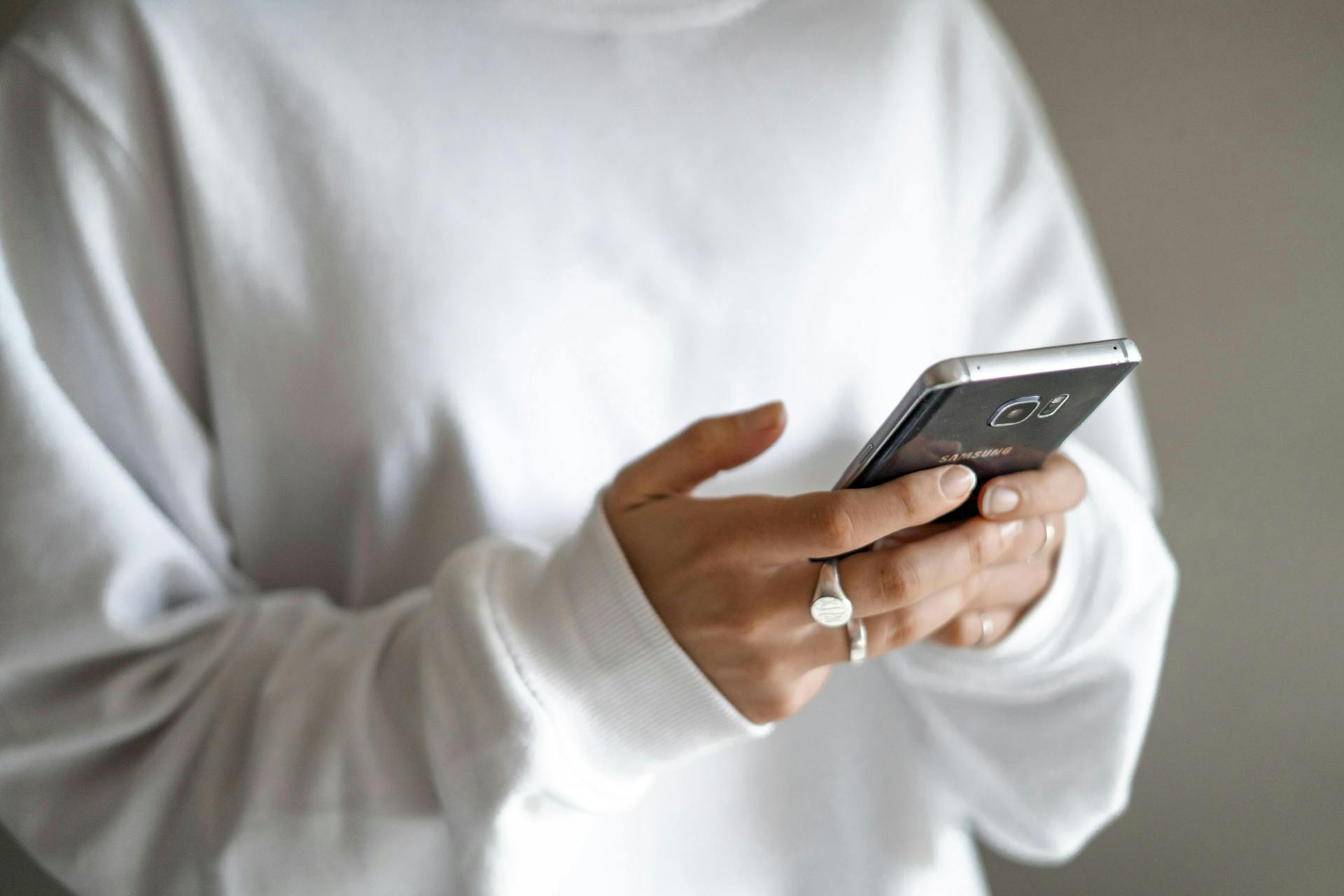 A person wearing a white long-sleeved shirt uses a Samsung smartphone, typing or browsing, in a neutral, dimly lit space.