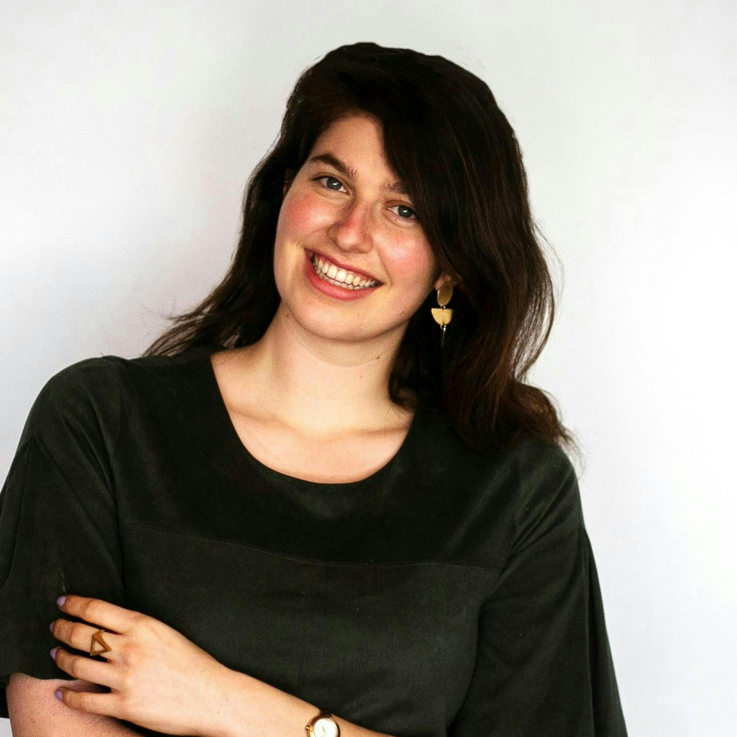 A woman wearing a dark green top and gold earrings smiles while crossing her arms against a plain, light-colored background.