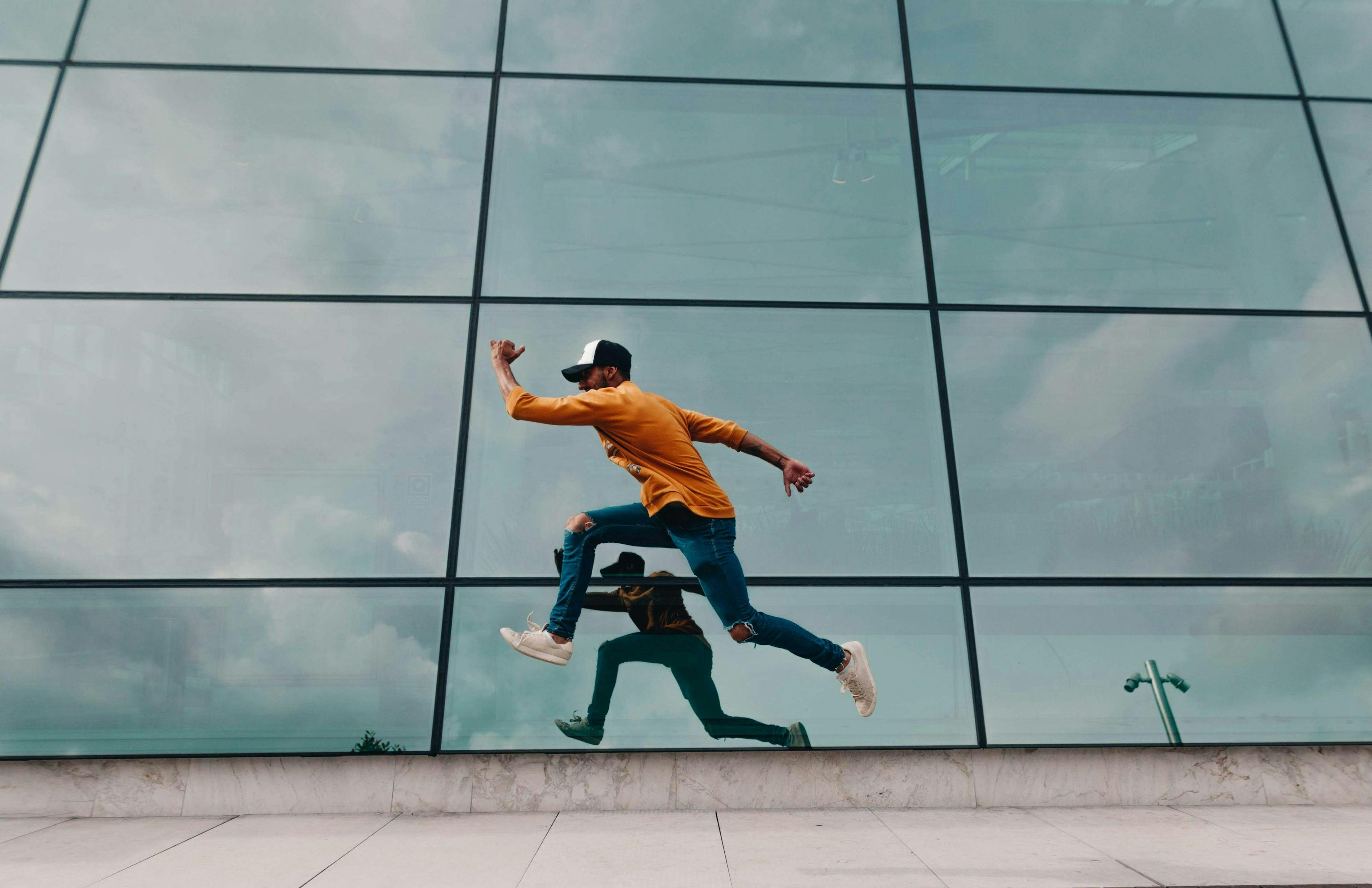 A person in a yellow sweater and cap leaps mid-air, reflected in a large glass building facade, against a cloudy sky backdrop.