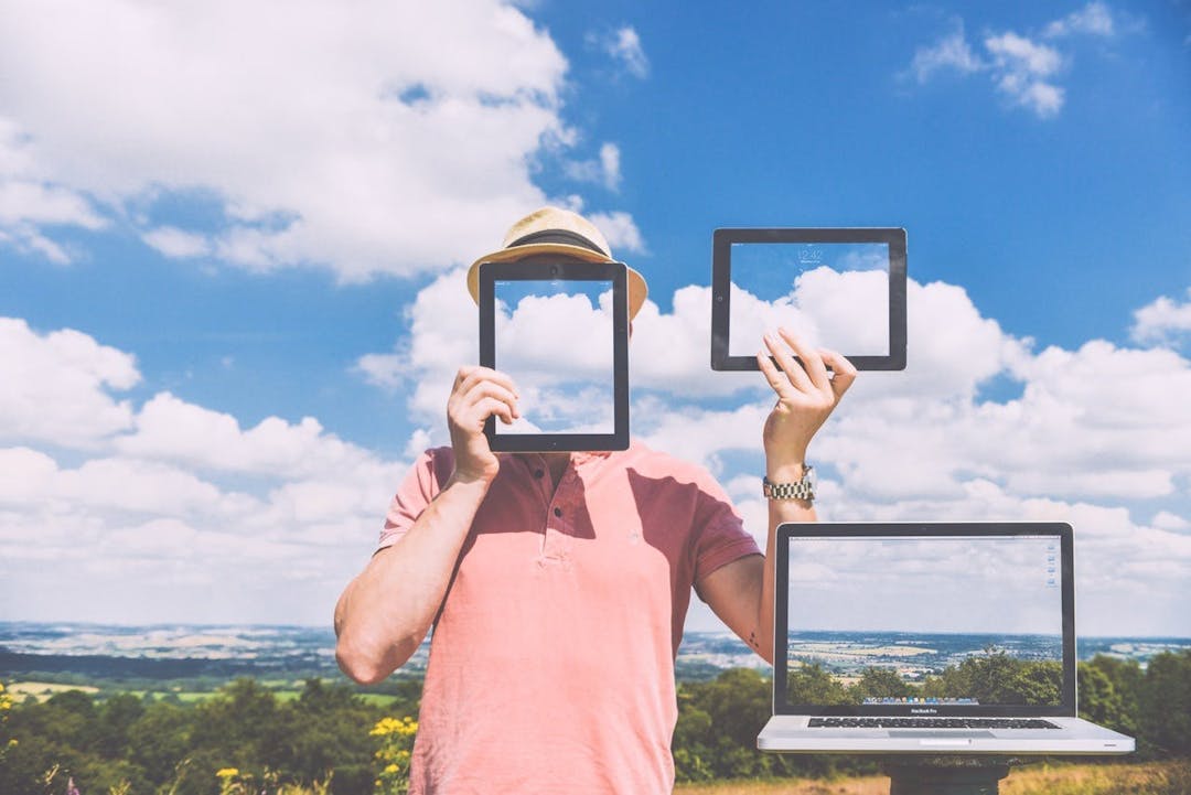 Person holding two frames against their face and body, with a laptop on a stand, all displaying a clear sky, standing in a field under a bright, cloudy sky.