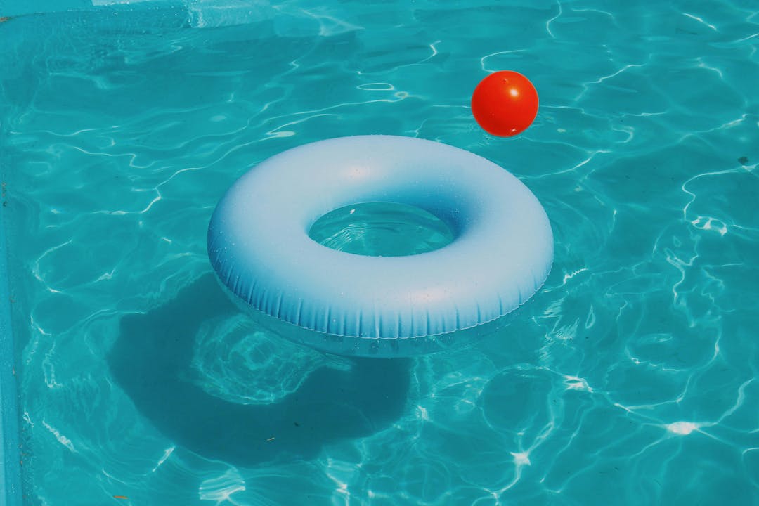 A blue inflatable ring floats in a clear, sunlit swimming pool with rippling water. An orange ball hovers nearby.