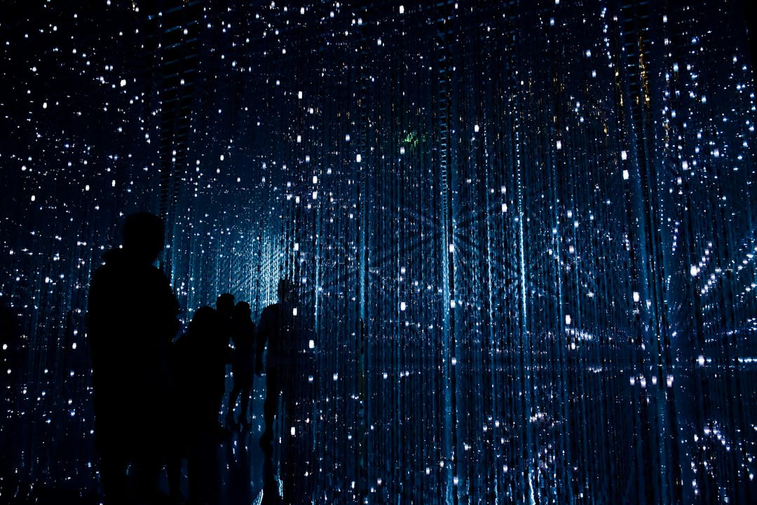 Silhouetted figures stand, observing light strands suspended in a dark, immersive room resembling a starry sky, creating an ethereal, almost cosmic atmosphere.