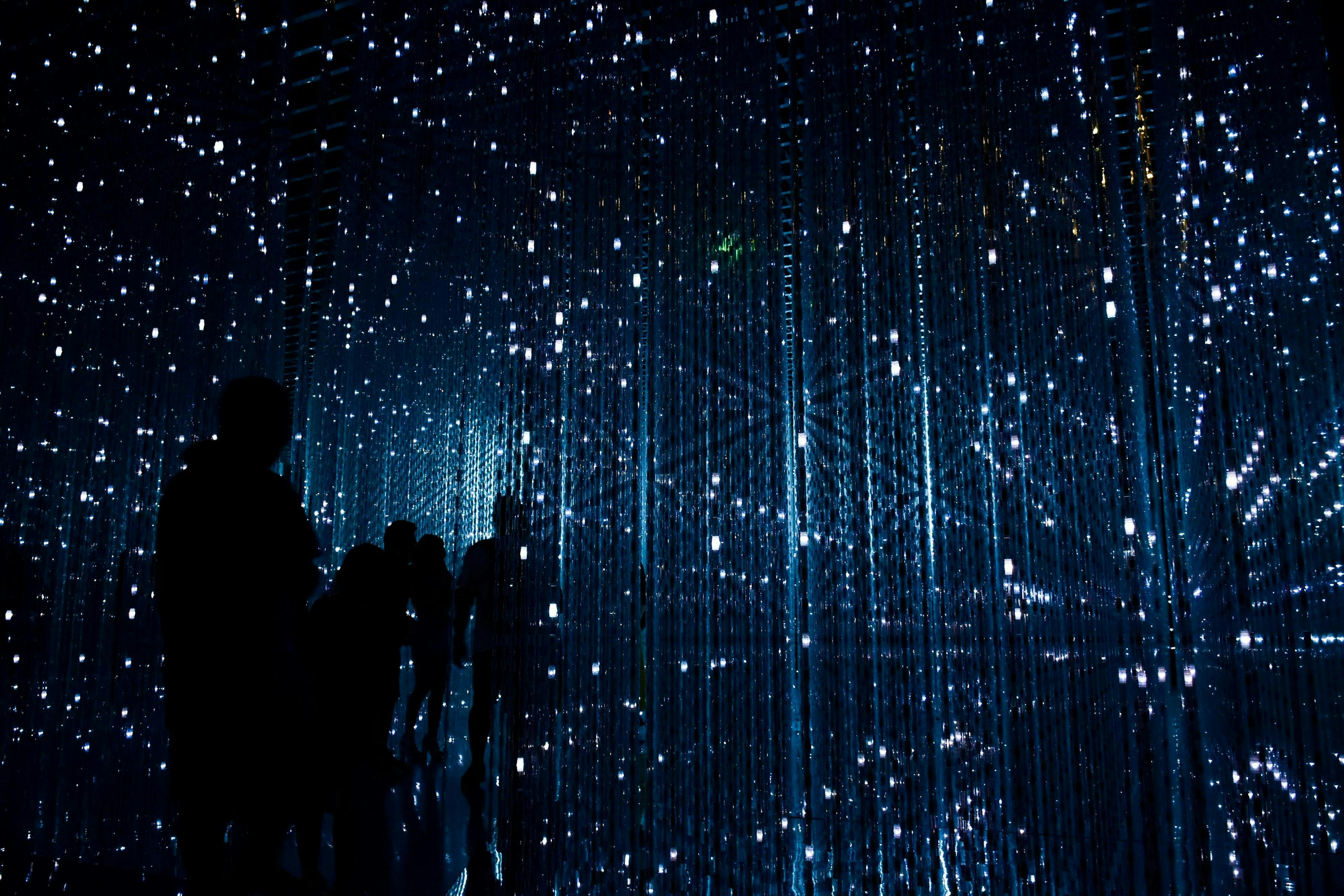 Silhouetted figures stand, observing light strands suspended in a dark, immersive room resembling a starry sky, creating an ethereal, almost cosmic atmosphere.