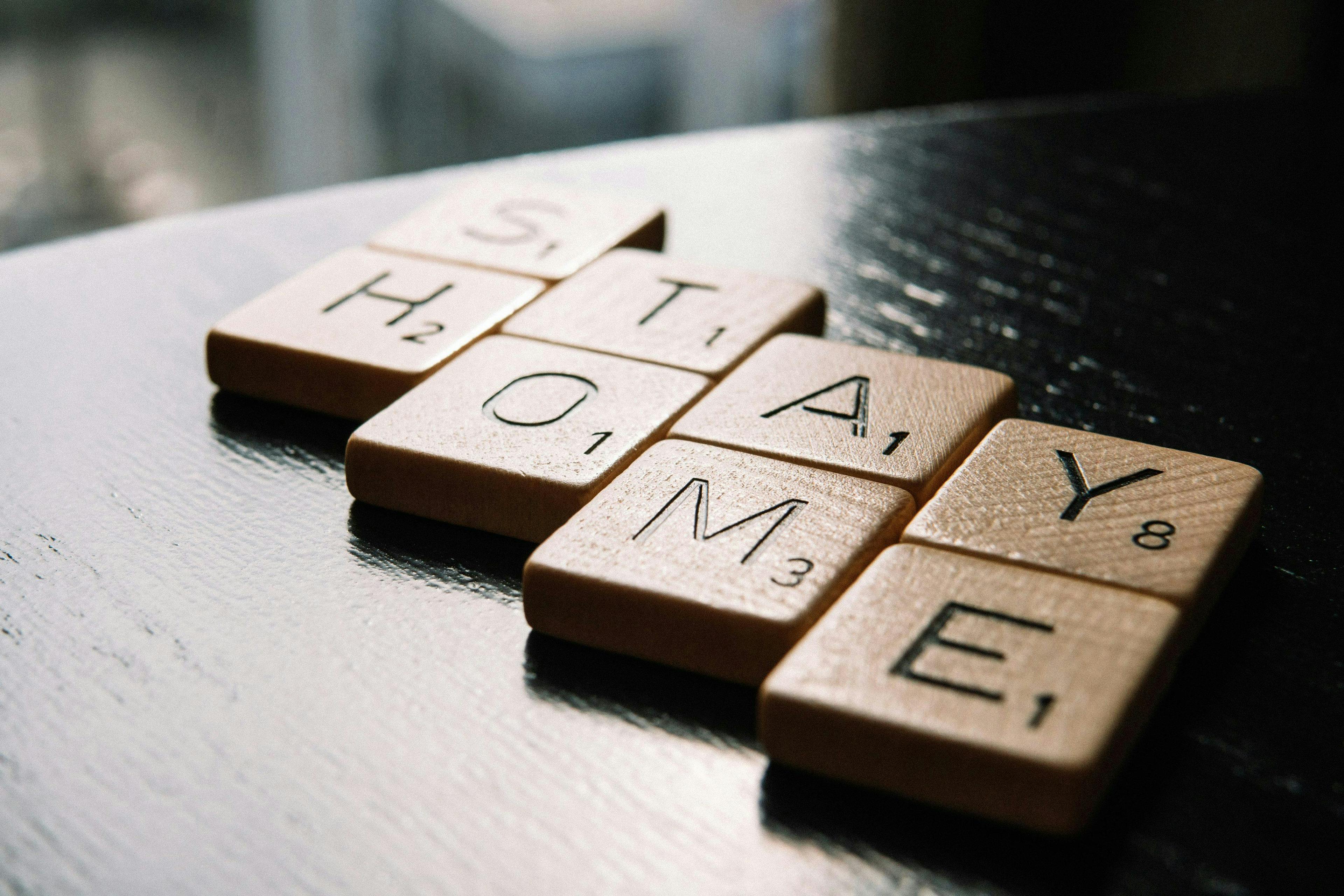 Wooden letter tiles spell "STAY HOME" on a dark wooden surface, with natural light coming through a blurred indoor background.