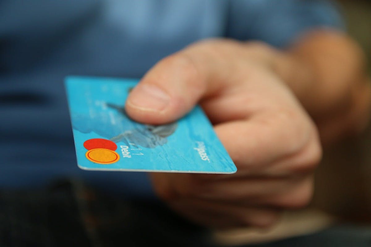 A hand holds and extends a blue debit card with visible Mastercard logo and the text "debit" and "spark" in a blurred background, indicating a payment action.