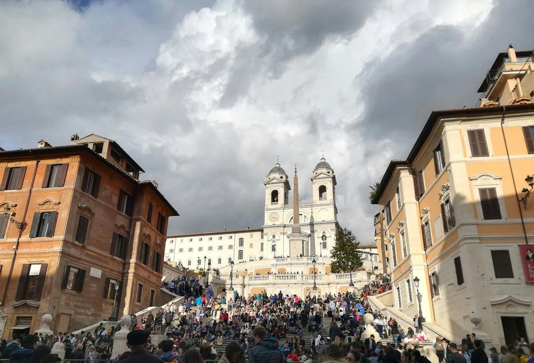 People are sitting and walking on the Spanish Steps, flanked by historic buildings, leading up to the Trinità dei Monti church, under a cloudy sky.