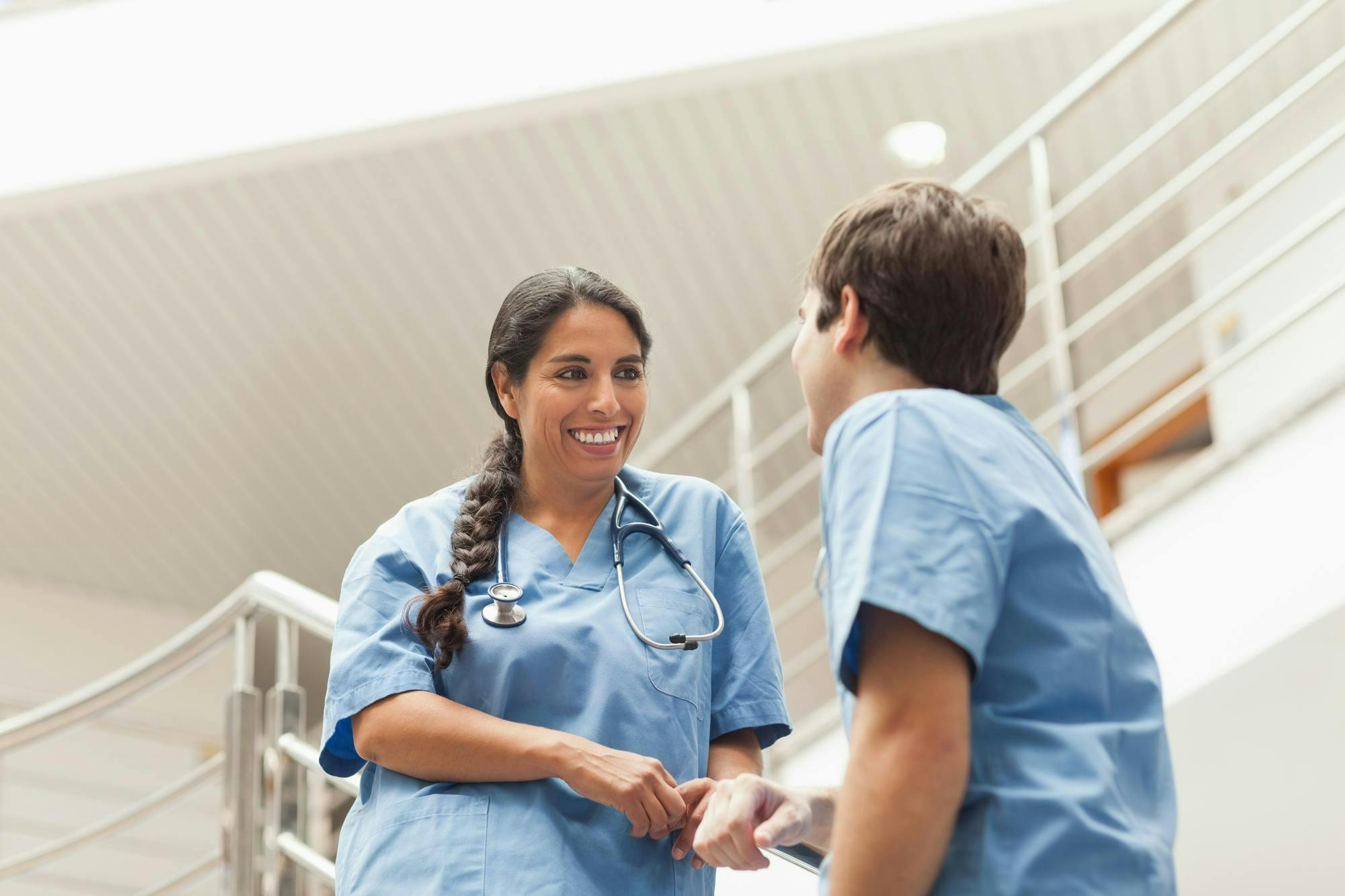Two healthcare professionals in blue scrubs chatting amiably near a staircase in a medical facility; one has a stethoscope around her neck and both appear engaged in conversation.
