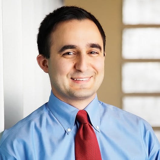 A man is smiling while wearing a light blue dress shirt and red tie in an indoor setting with a blurred window background.