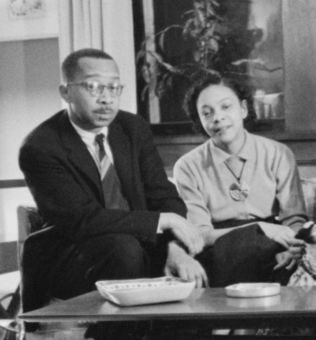 A man in a suit and a woman in a dress sit side by side on a couch in a domestic setting, with a table featuring a dish and ashtray in front of them.