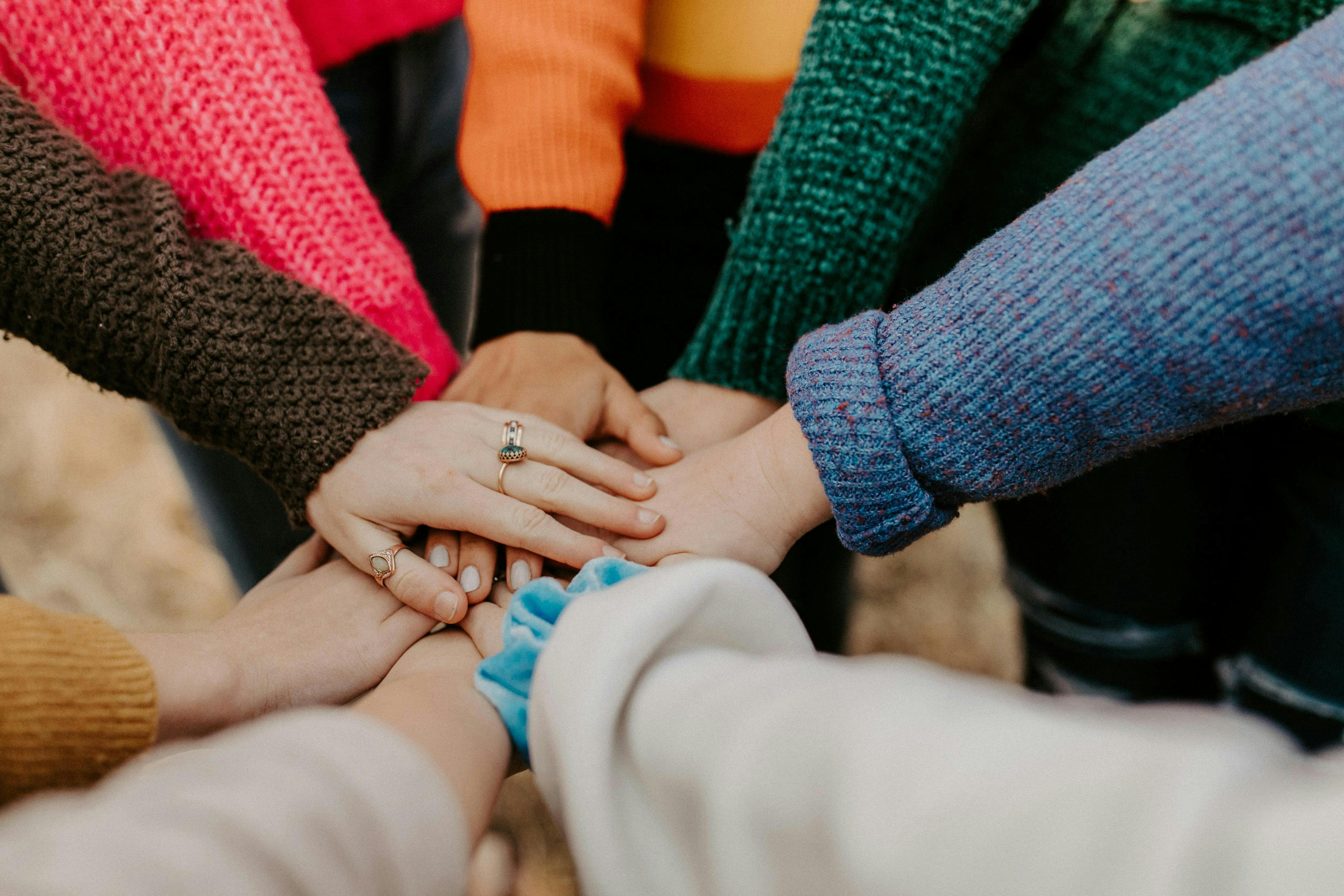 A group of people, wearing colorful sweaters, place their hands together in the center, signifying unity and teamwork in an outdoor setting.