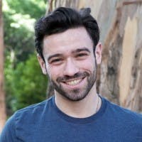 A man with dark hair and a beard is smiling outdoors, wearing a blue shirt against a backdrop of green foliage and a wooden fence. No text is present.