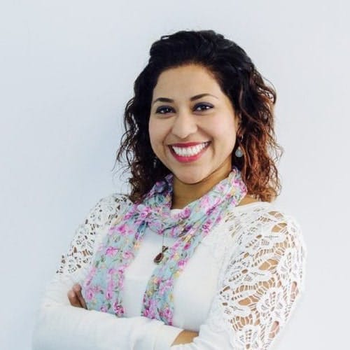 A woman with curly hair is smiling, arms crossed, wearing a white lace top and floral scarf, standing against a plain white background.
