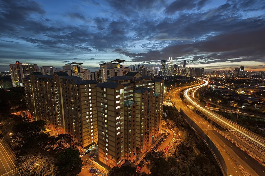 Tall apartment buildings with illuminated windows stand prominently in the foreground, while a winding highway with light trails indicates ongoing traffic; set against a backdrop of a city skyline at dusk.