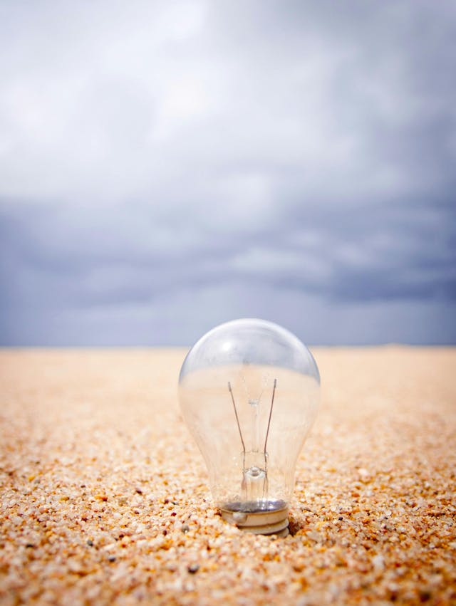 A light bulb stands upright, partially embedded in coarse sand, under an expansive, cloudy sky.
