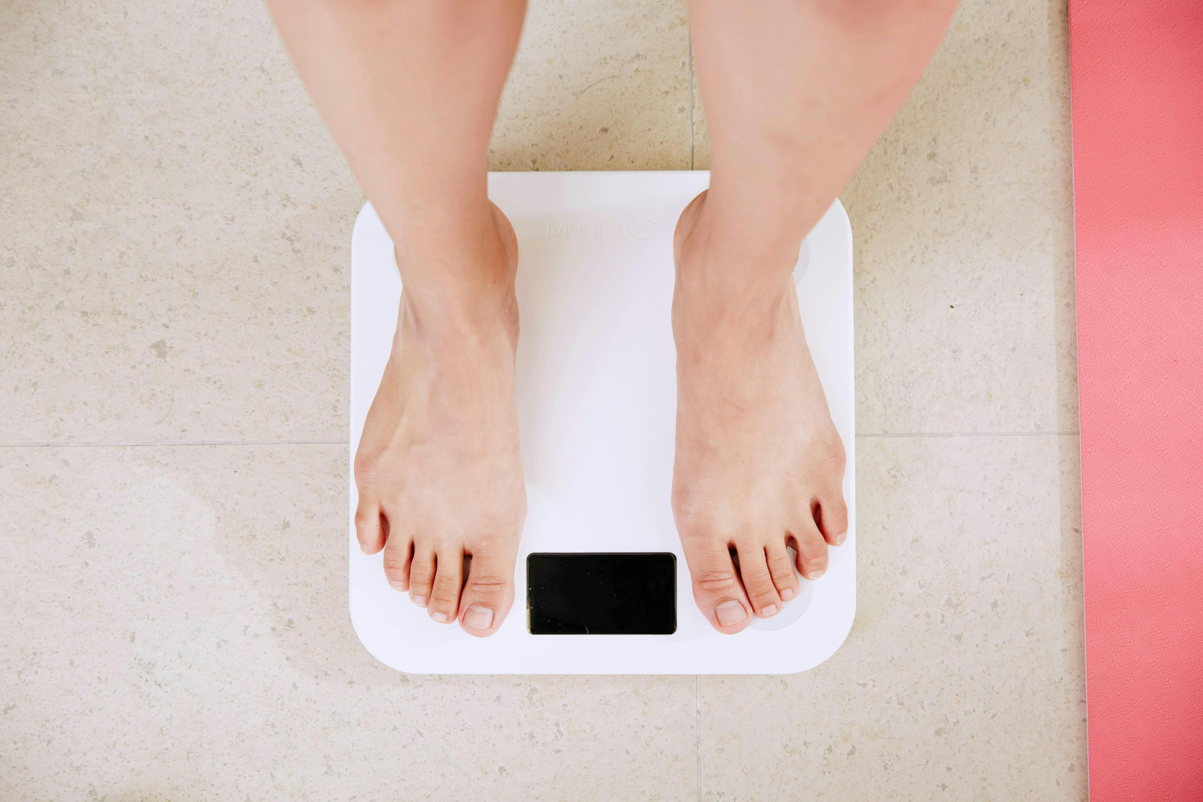 Feet standing on a white digital scale, with a pink yoga mat nearby, on a tiled floor.