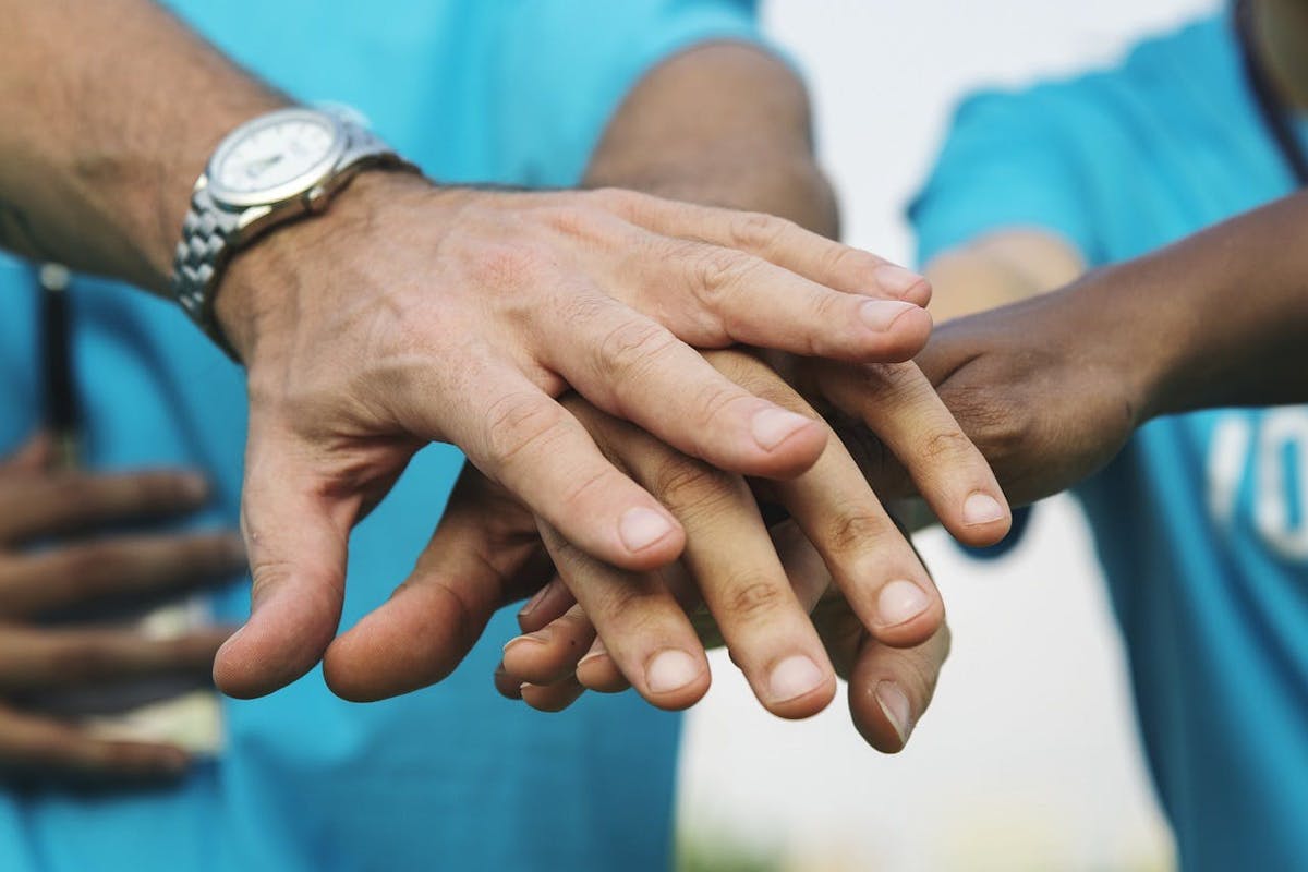 Hands placed together in a supportive gesture, with people in blue shirts in the background.