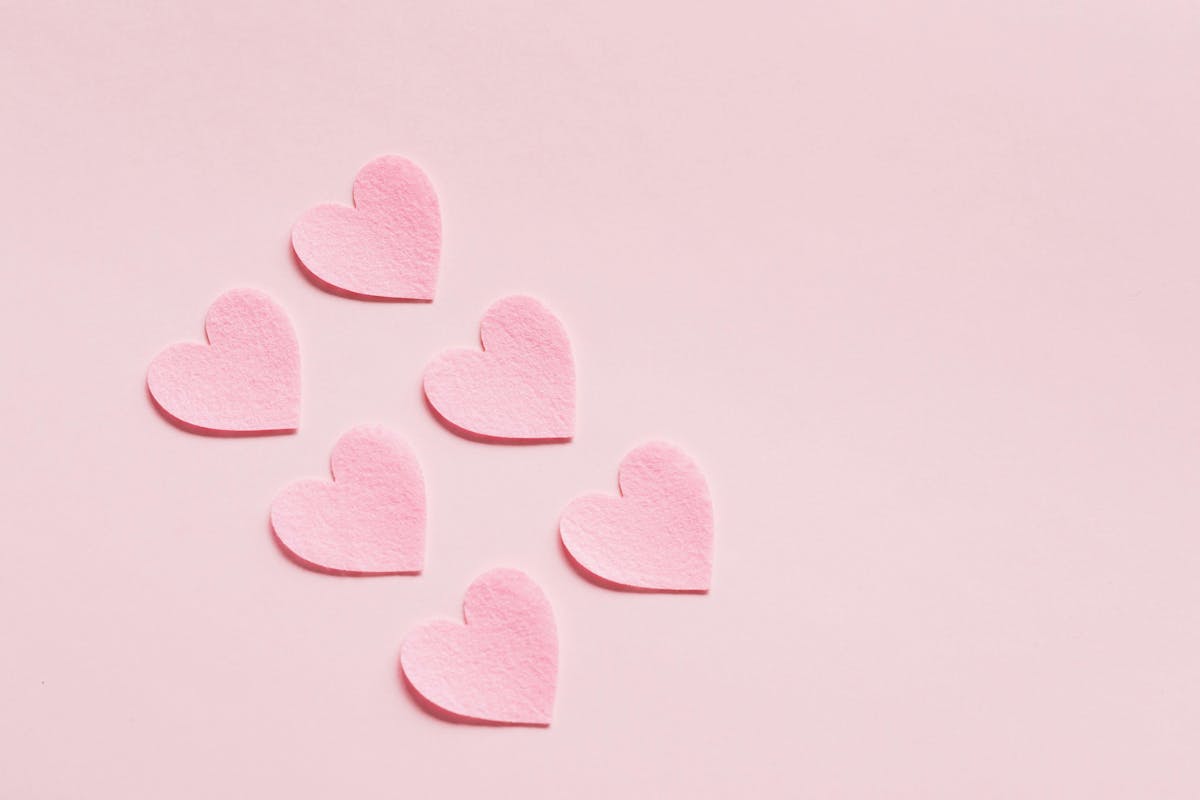 Six pink felt hearts lie scattered on a smooth, light-pink background, creating a simple yet charming design.