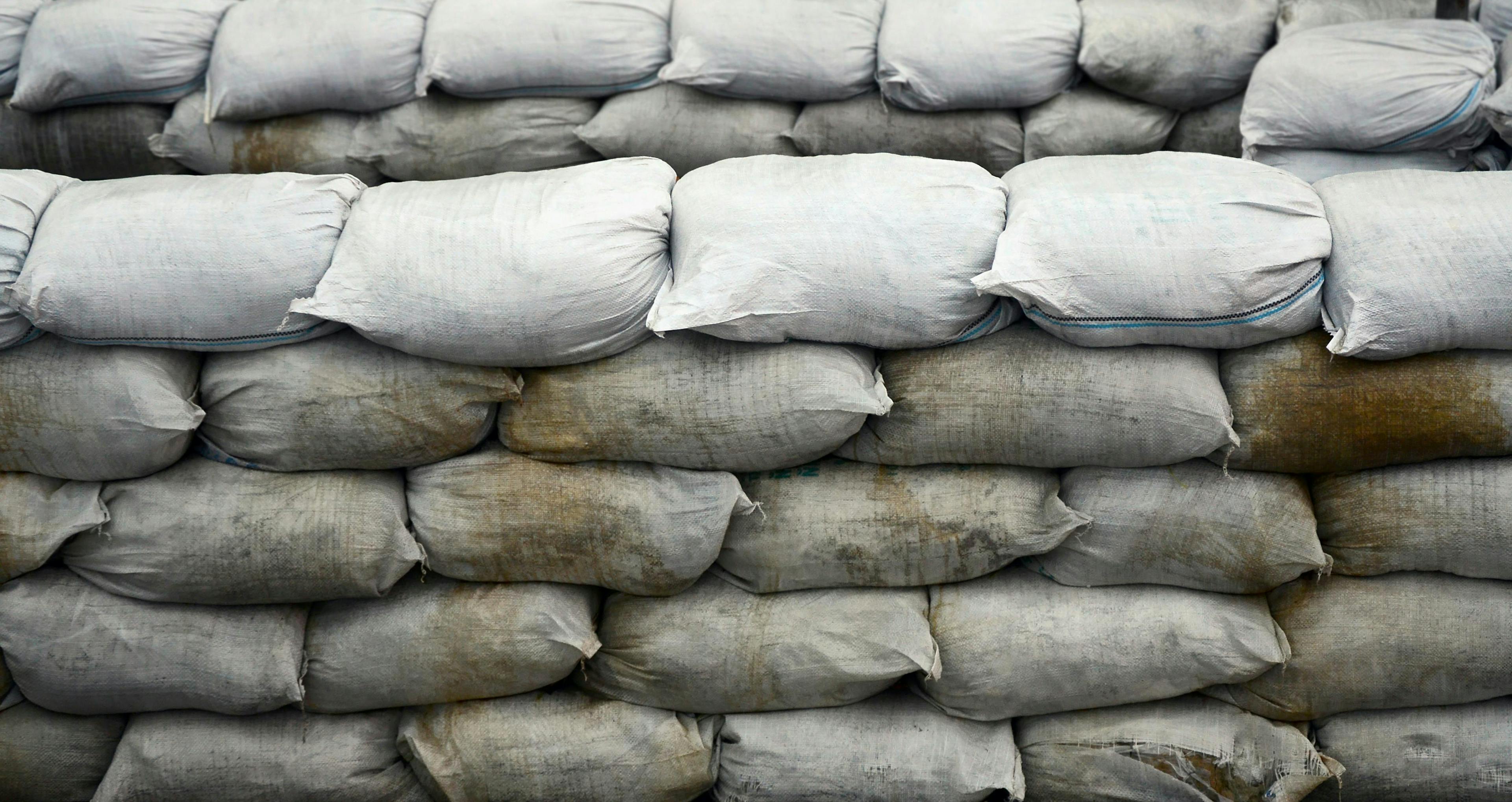 Stacks of grey sandbags, arranged in several horizontal rows, form a barricade. The sandbags are tightly packed and slightly weathered, indicating outdoor use, likely in a construction or defense setup.