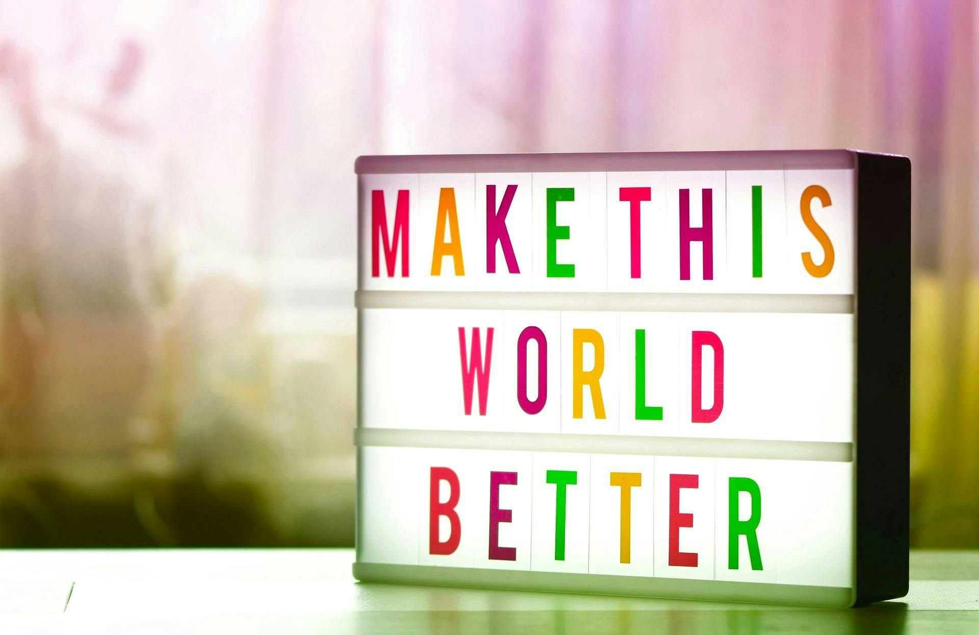 Lightbox sign displaying "MAKE THIS WORLD BETTER" in colorful letters, sitting on a table with a blurred light, warm-toned background.