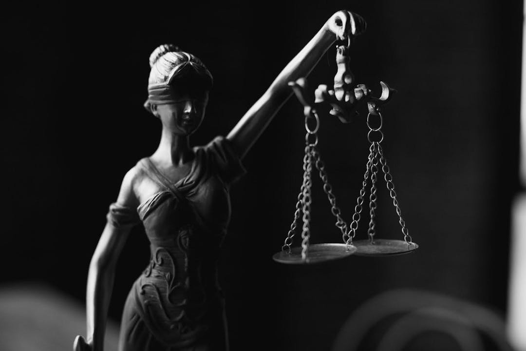 Blindfolded Lady Justice statue holds scales of justice; intricate gown details visible. The background is dark and out of focus, emphasizing the statue's symbolic form and balance.