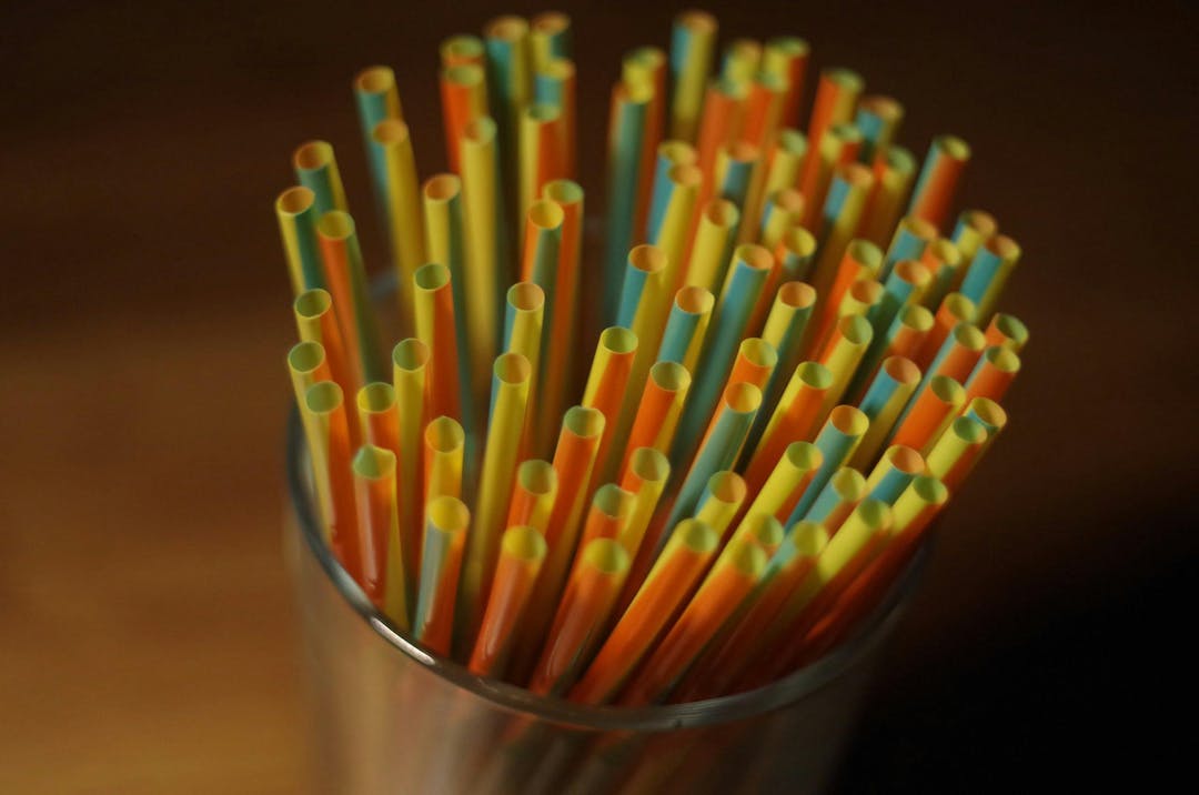 Multicolored plastic straws stand upright in a transparent glass on a wooden surface, arranged closely together with their open ends facing up, creating a vibrant pattern.