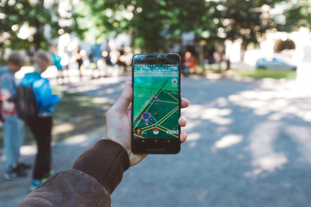 A hand holds a smartphone displaying a game app in an outdoor park area with blurred people and trees in the background.
