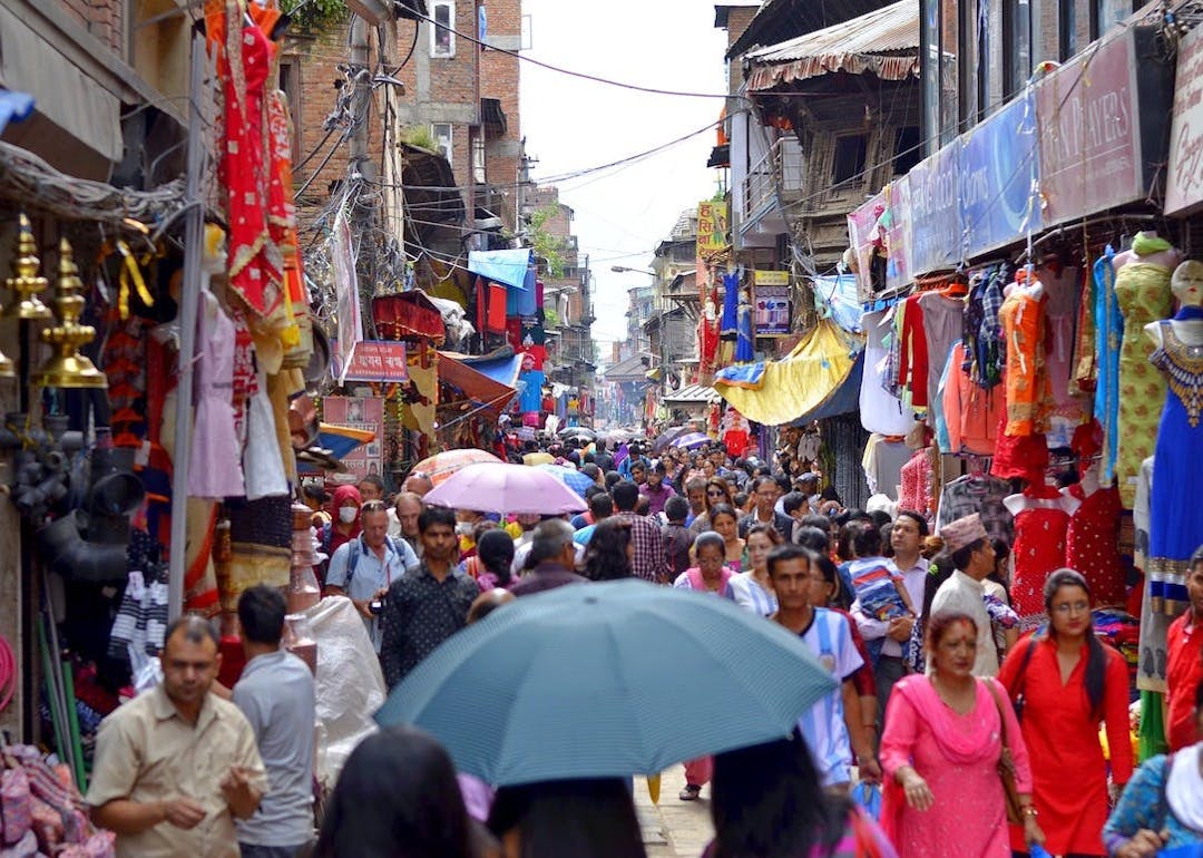 Crowds of people walk through a bustling, narrow market street flanked by colorful textiles and goods. An individual holds an umbrella in the foreground amidst vibrant market stalls and shops.