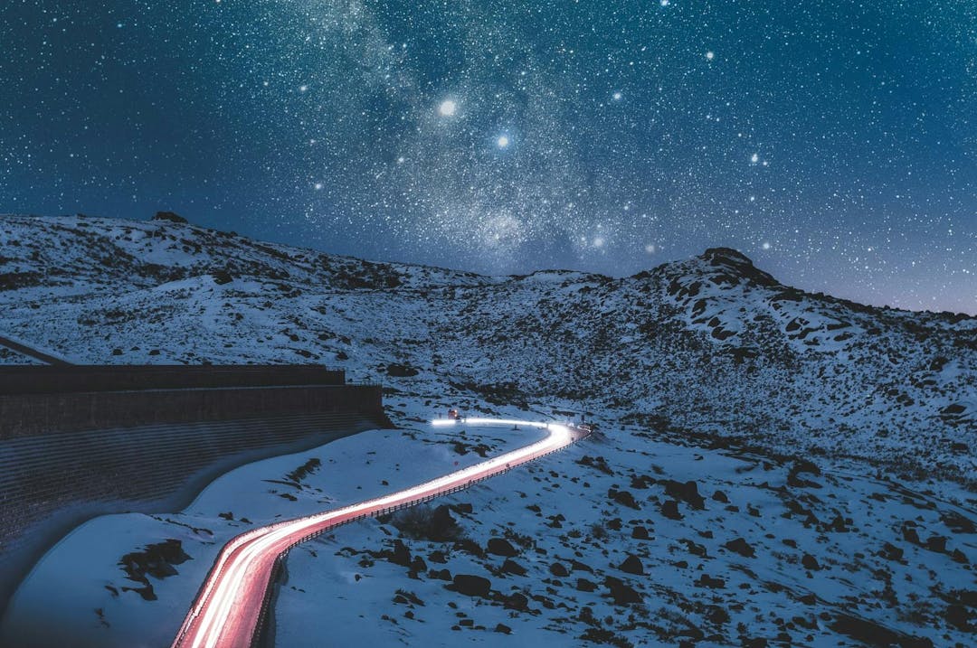 A winding road, illuminated by car lights, cuts through a snowy, mountainous landscape under a vast, star-filled night sky.