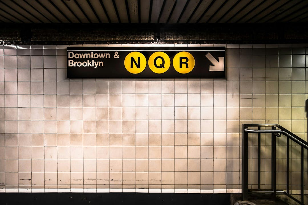 Sign for Downtown & Brooklyn N, Q, R subway lines, with a right arrow, mounted on a tiled wall above a staircase in a subway station.